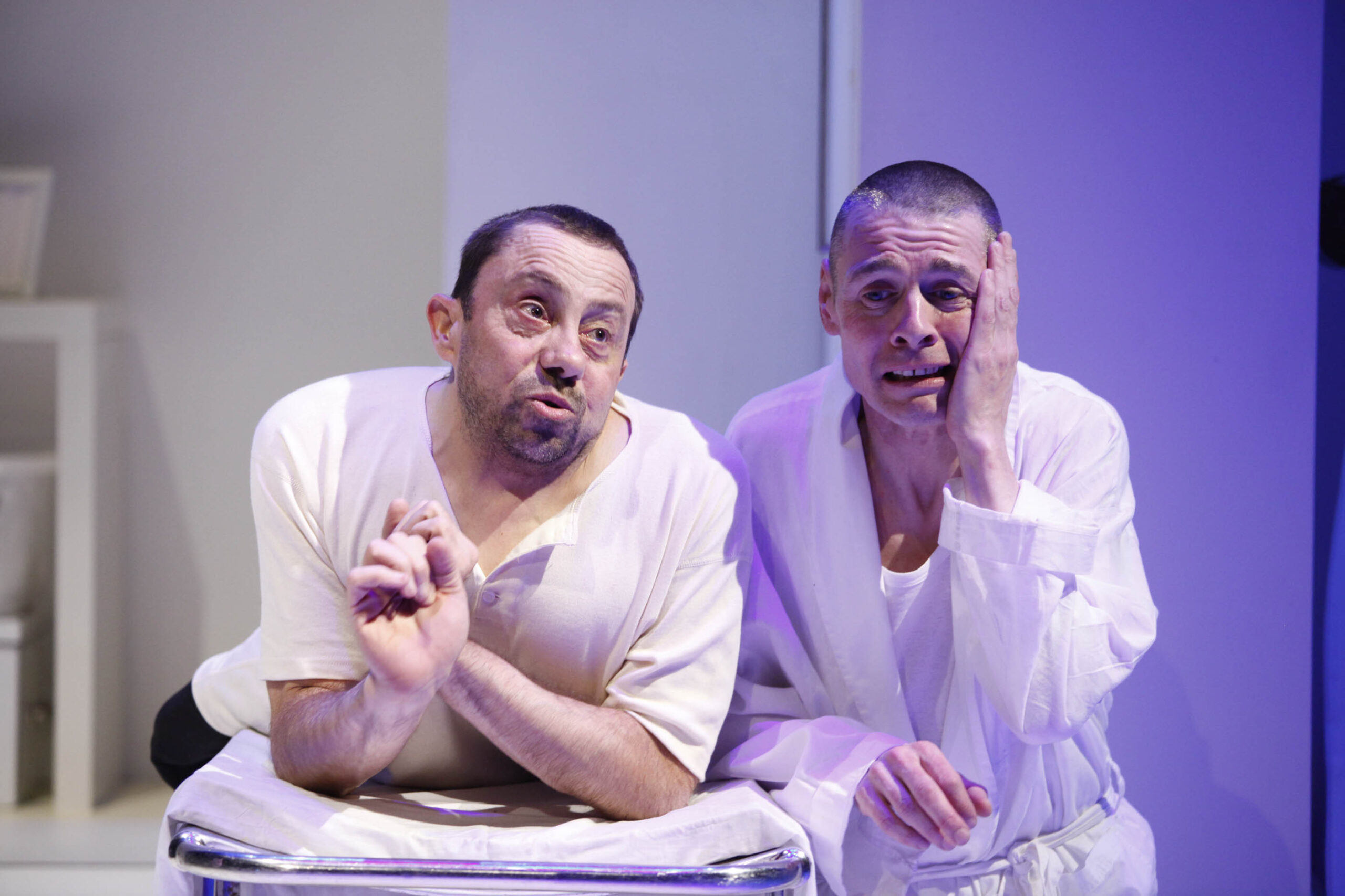 Two middle aged men leaning on a table. The one on the left is wearing a white shirt and the one of the right is wearing a white robe, he is holding one of his hands to his face and looks distressed.