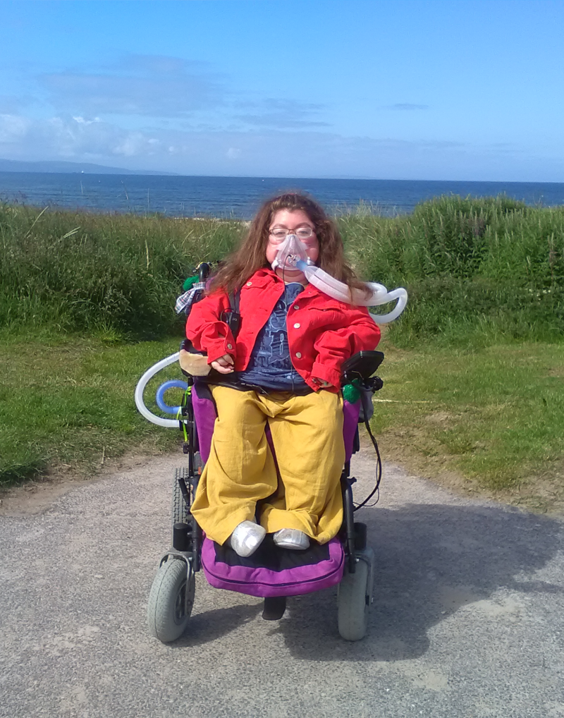 A young woman with brown hair, glasses and a medical mask, sitting in a pink wheelchair with her back to the ocean.