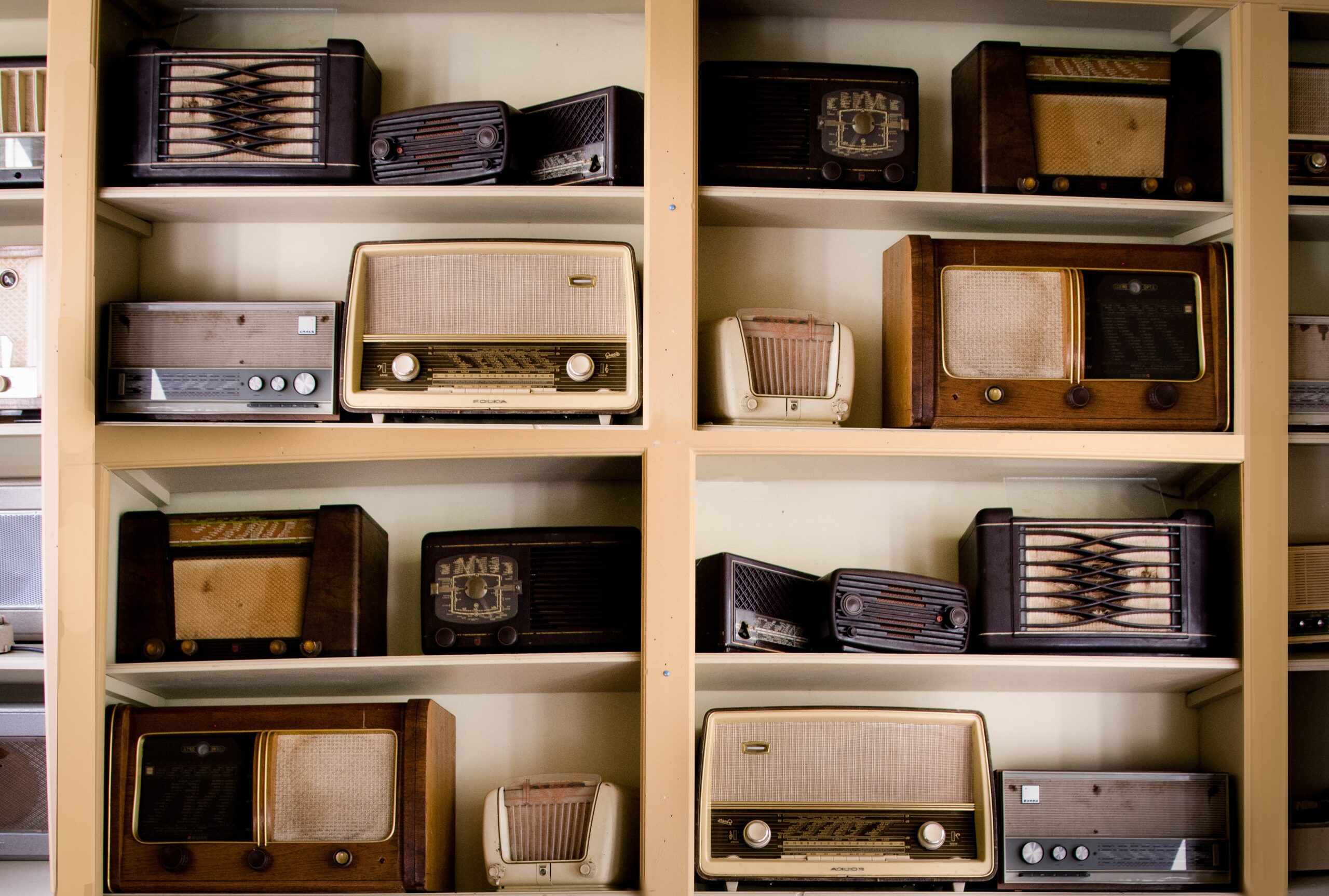 A set of shelves that are littered with lots of old style radios, all in different shades of muted browns some dark greys.