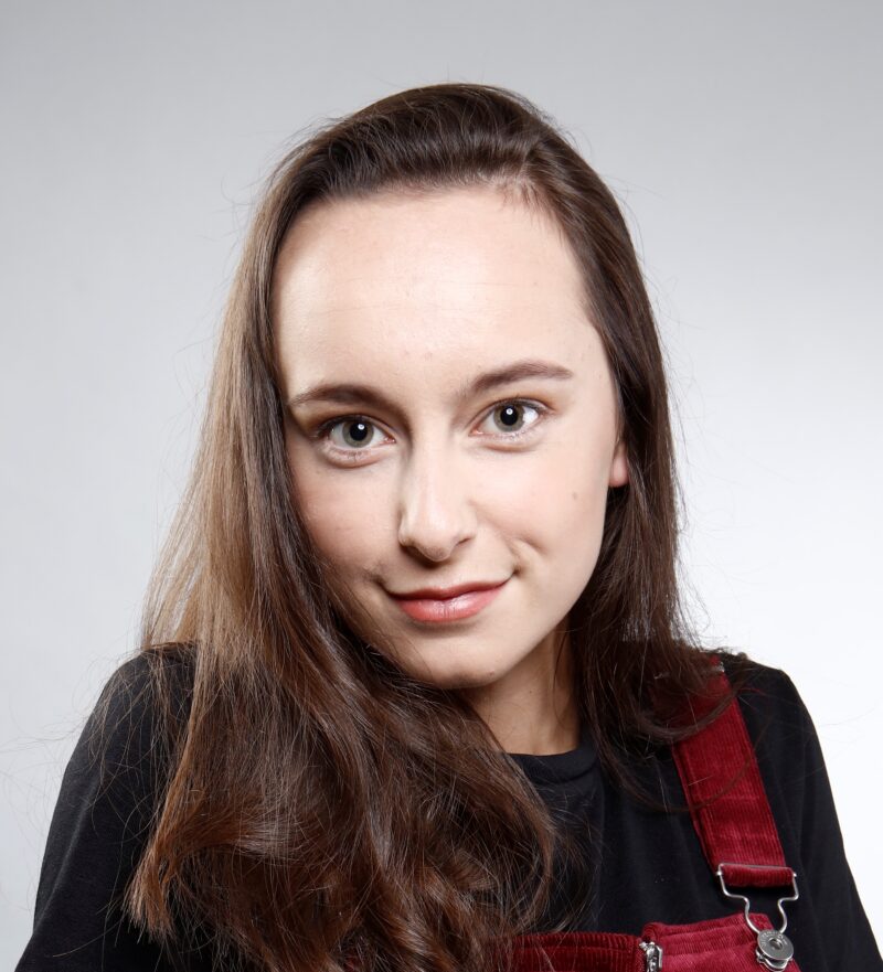 Katie anna is a white woman in her 20s. She has dark brown hair, partly tucked back behind one ear on one side, and partly cover her face on the other side. She is giving a slight, cheeky grin to the camera.